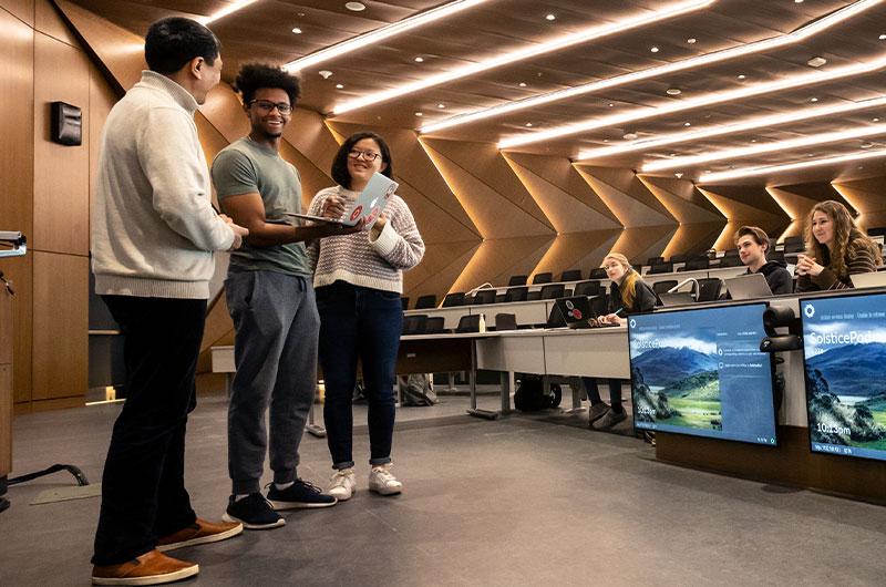 two students and an instructor in the front of a lecture hall looking at a laptop that one of the students is holding