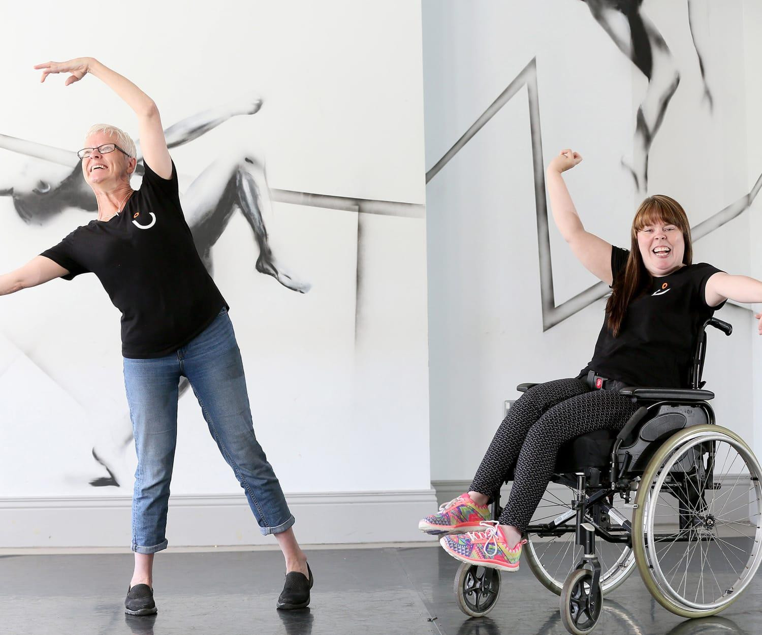 Two disabled dancers, one a wheelchair user, perform in a dance studio, with expressive drawings of dancers on the walls behind