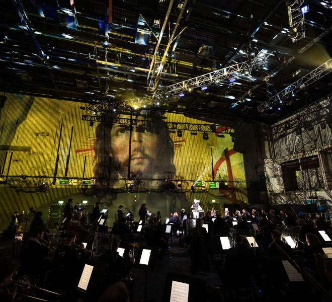 Orchestra perform in vast auditorium against backdrop projection of painting of face of Jesus Christ