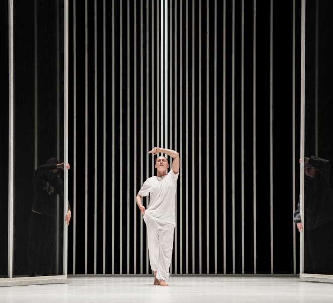 Contemporary dancer dressed all in white performs barefooted on stage against black and white striped backdrop.