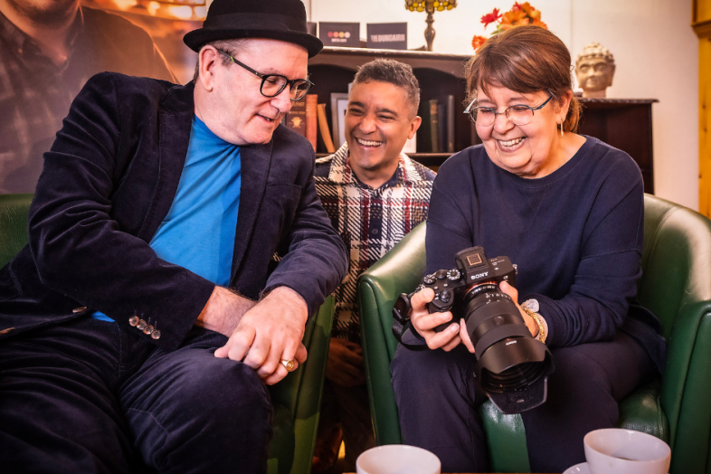 Group of three people smiling and looking at a camera. Two are seated and one is kneeling behind.