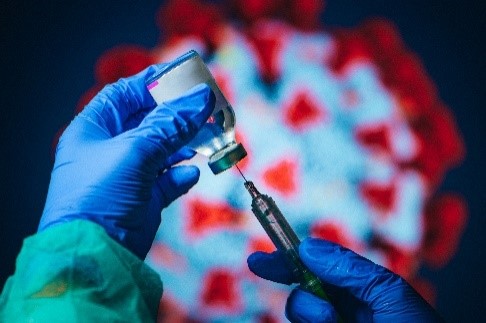 Hands in medical gloves drawing liquid from a vial into a syringe with a model of SARS-CoV-2 in the background.