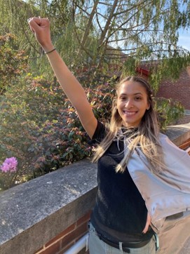 Keishla Marrero Acosta standing in front of green plants, with a silver cape and one arm raised in a superhero pose.