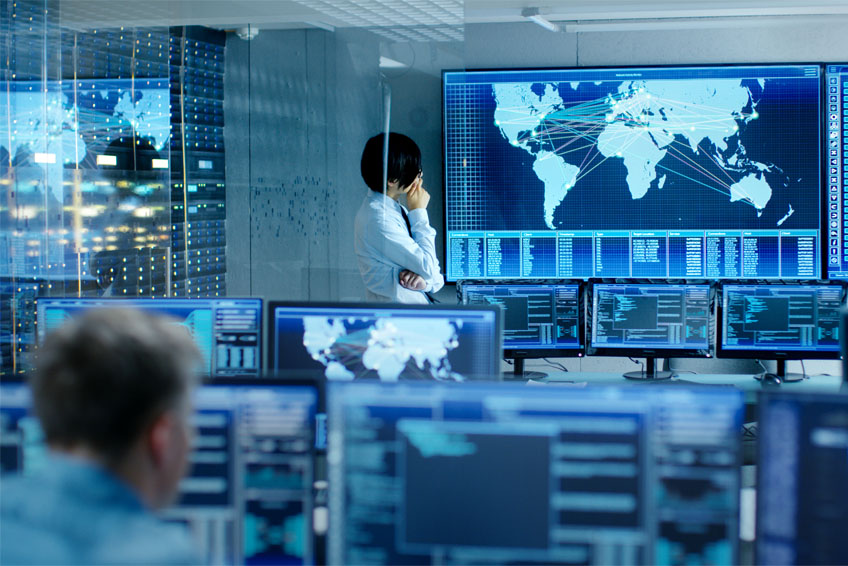 A person in a security operations center stands in front of a large monitor with a map of the world in a room with multiple monitors displaying security data