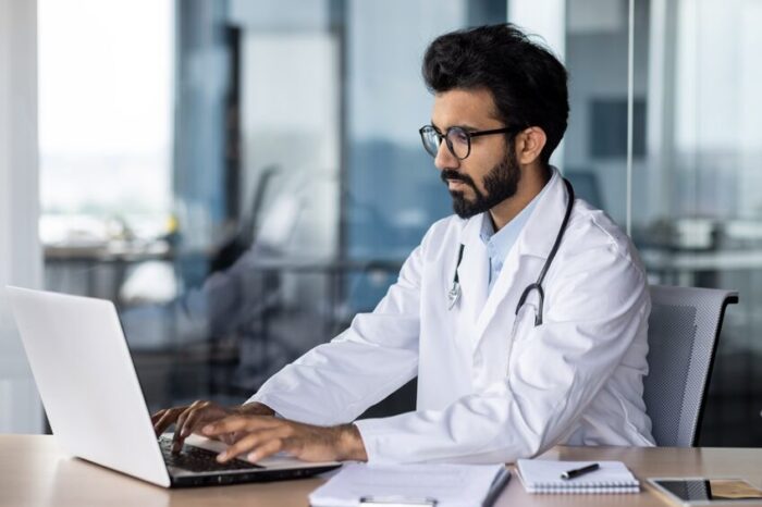 A doctor wearing a lab coat with a stethoscope works at a laptop