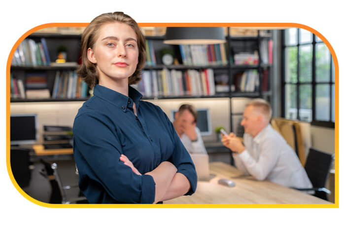 A professional with crossed arms stands confidently in an office space with bookshelves and two people sitting at a shared desk in the background