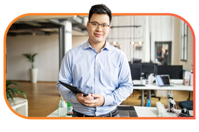 A person in business casual attire and glasses holds a tablet device in an office setting and looks at the camera