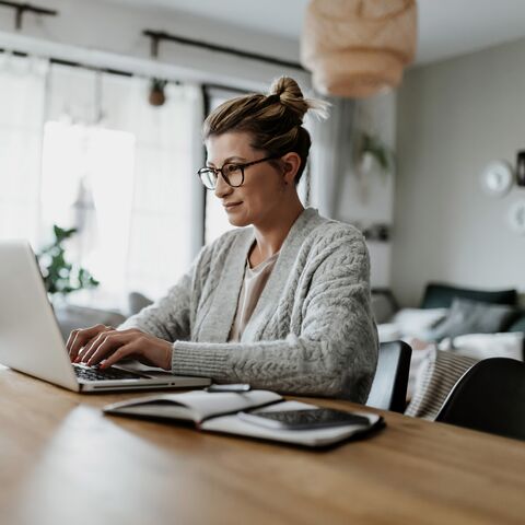 A woman with glasses and her hair up is working intently on a laptop in a bright, cozy living room with a coffee cup.