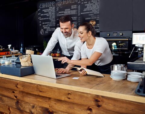 Un uomo e una donna in un caffè sorridono e lavorano insieme a un computer portatile, circondati da utensili da cucina e da un menu sullo sfondo.