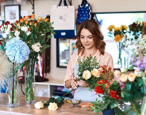 A florist with a striped blouse arranges various bouquets of flowers on a wooden table in a light-colored flower store.