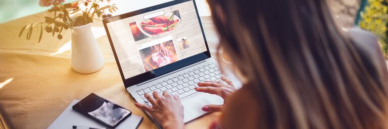 Une femme est assise à une table et tape sur un ordinateur portable. Un carnet de notes, un stylo et un smartphone sont posés à côté d'elle. Un vase rempli de fleurs est posé sur la table.