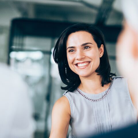 Une femme souriante aux cheveux noirs et au chemisier gris serrant la main de quelqu’un d’autre au premier plan.