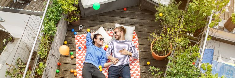 Un couple est allongé en riant sur une terrasse de toit, sur une couverture à motifs rouges et blancs, entouré de plantes et de balles multicolores.