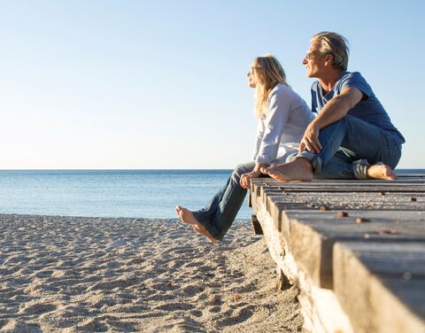 Ein Mann und eine Frau sitzen entspannt auf einem Holzsteg am Strand und blicken lächelnd aufs Meer unter klarem, blauem Himmel.