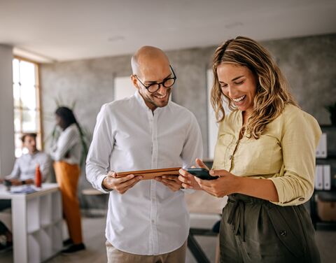 Two smiling office workers, a man with a tablet and a woman with a smartphone, are standing in a modern office discussing something.