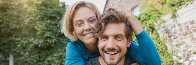 Un couple souriant, la femme enlace l'homme par derrière, tous deux portent des hauts bleus, à l'arrière-plan se trouve un paysage de jardin vert.