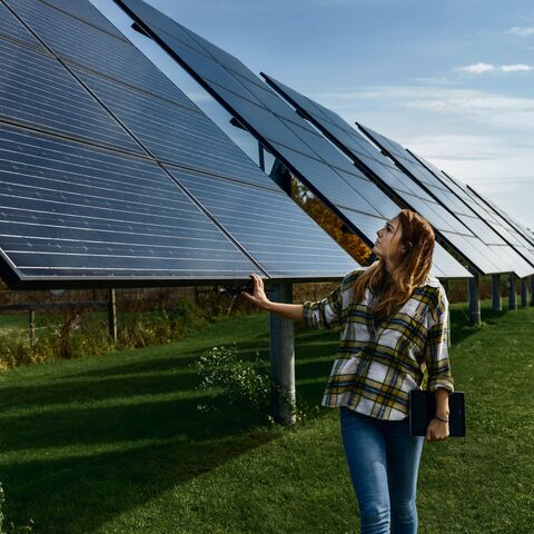 Une femme en chemise à carreaux et en jean inspecte à l’extérieur une série de grands panneaux solaires par un temps ensoleillé et tient une tablette à la main.