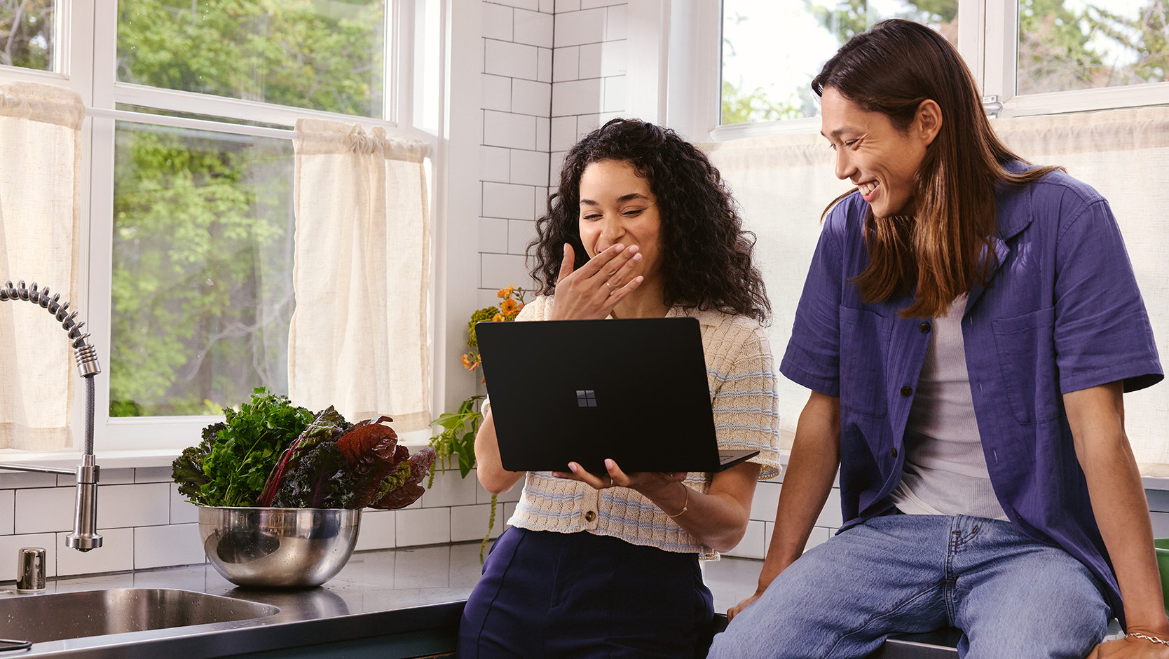 Two friends use a laptop in a kitchen.