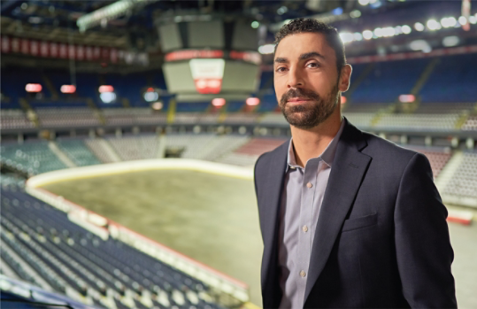 Ziad Mehio staat in een stadion en draagt een marineblauwe sportjas.