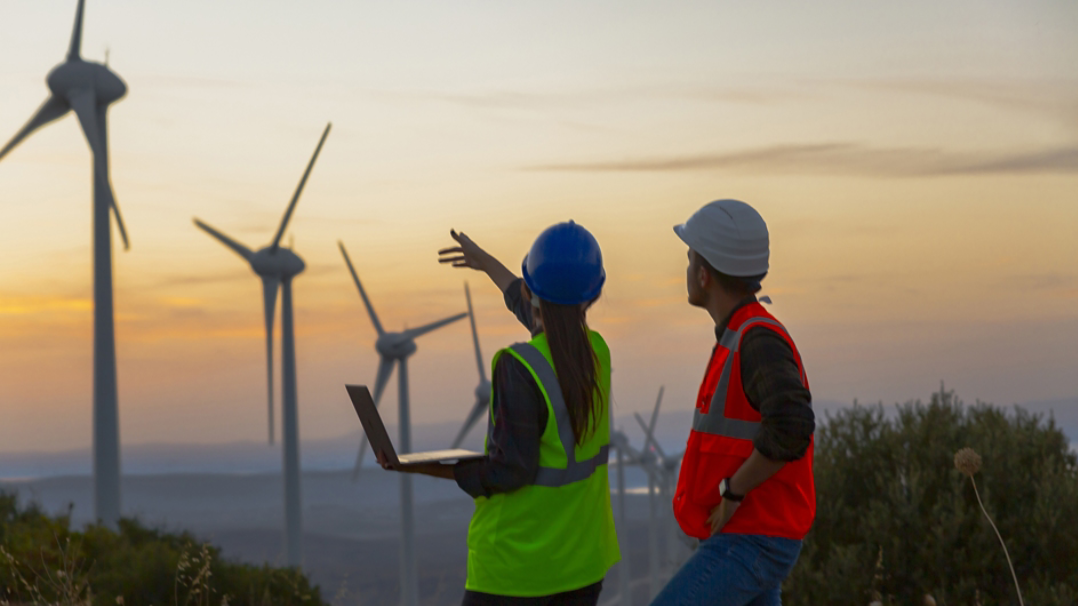 Two workers look out over a wind energy field while one holds a laptop.