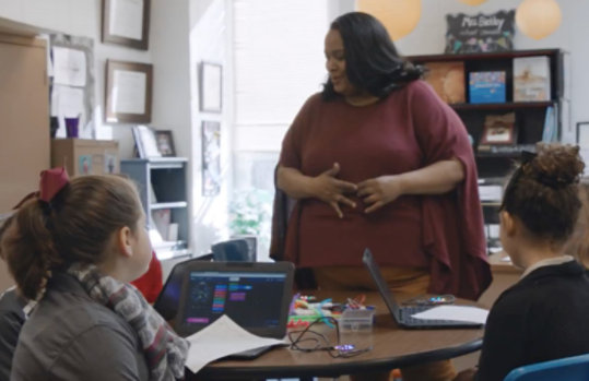 A teacher addresses students sitting at a table with laptops in front of them.