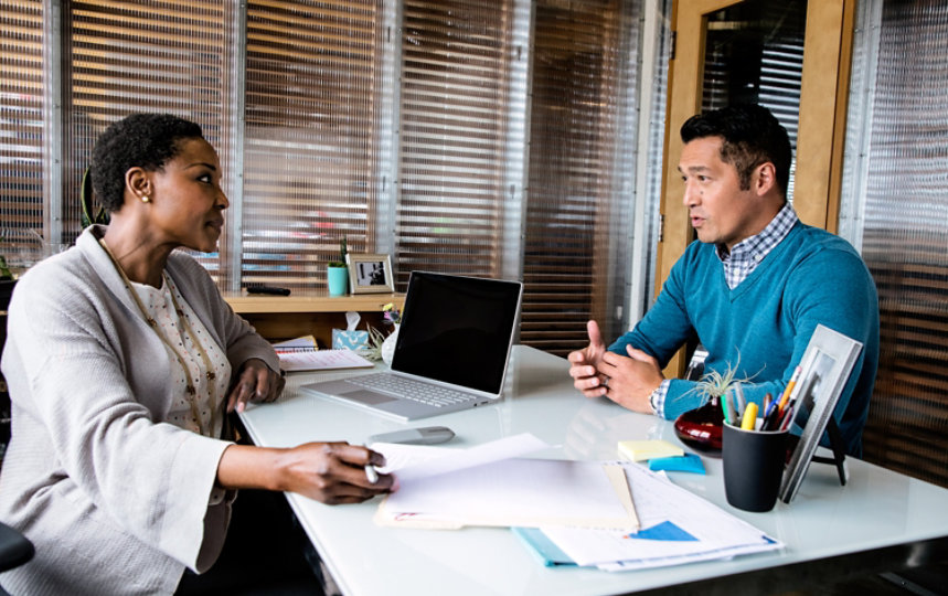 A woman and a man talking in her office with a laptop on the desk.