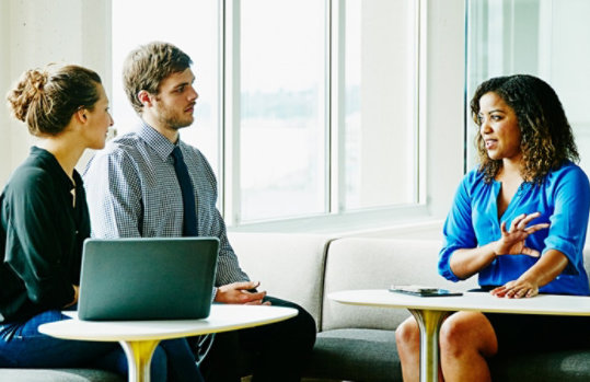 A businesswoman leads a meeting with two colleagues in a sunny office.