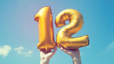 A gold foil number 12 balloon is held high in the air by male hands; photo taken outdoors on a bright sunny day, the sky is blue with some clouds. A vintage style effects has been added to the image.