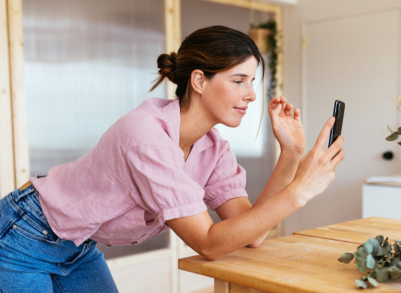 Young woman wearing a pink shirt, leaning over a kitchen island, looking and smiling slightly at her phone