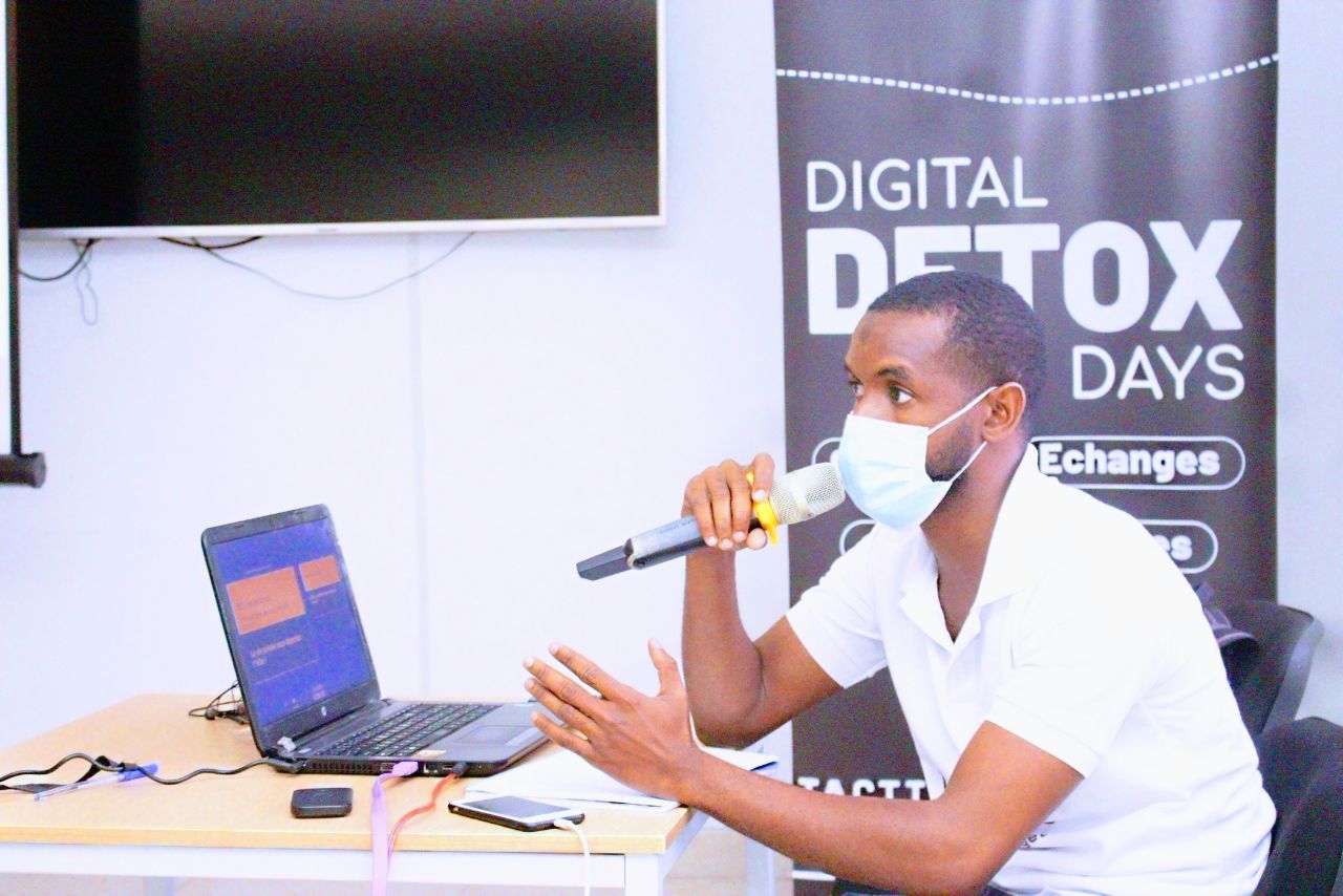 Photo of trainer with a microphone, seated in front of a computer, in front of the sign for the Digital Detox Days