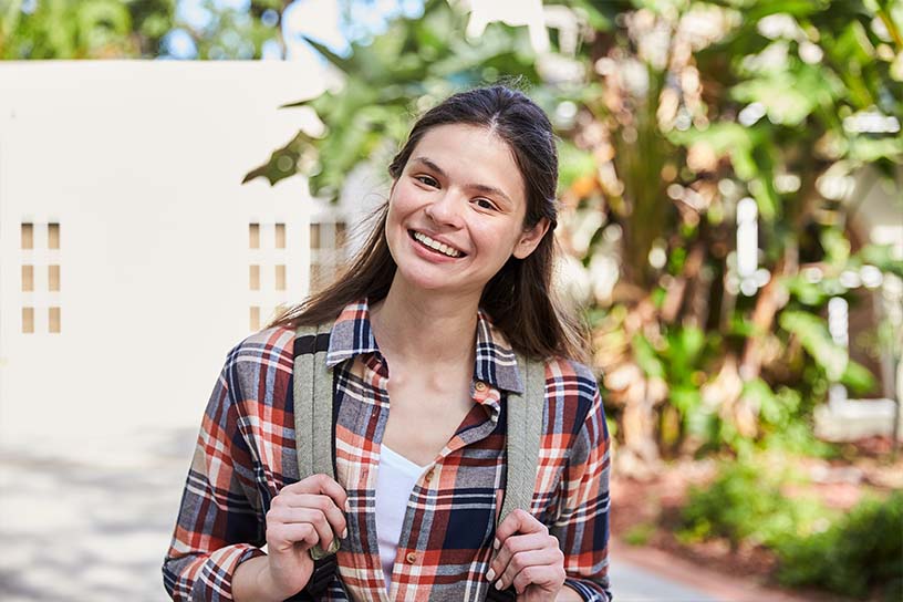 A young woman standing outside wearing a gray backpack, smiling toward the camera.