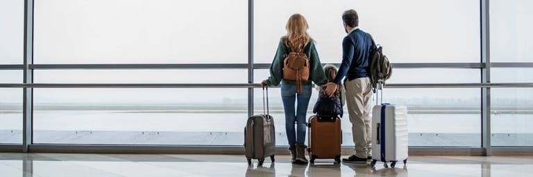 family of three with backpacks and carry-on bags looking out of the airport windows