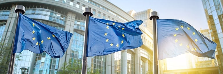 Three blue EU flags in front of a building with glass windows