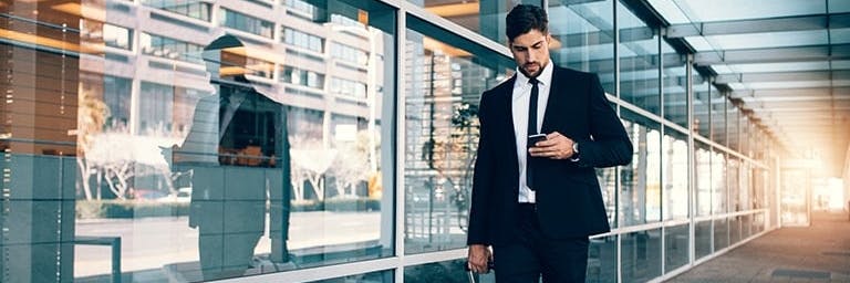 man in black suit walking with a carry-on bag and looking at his phone