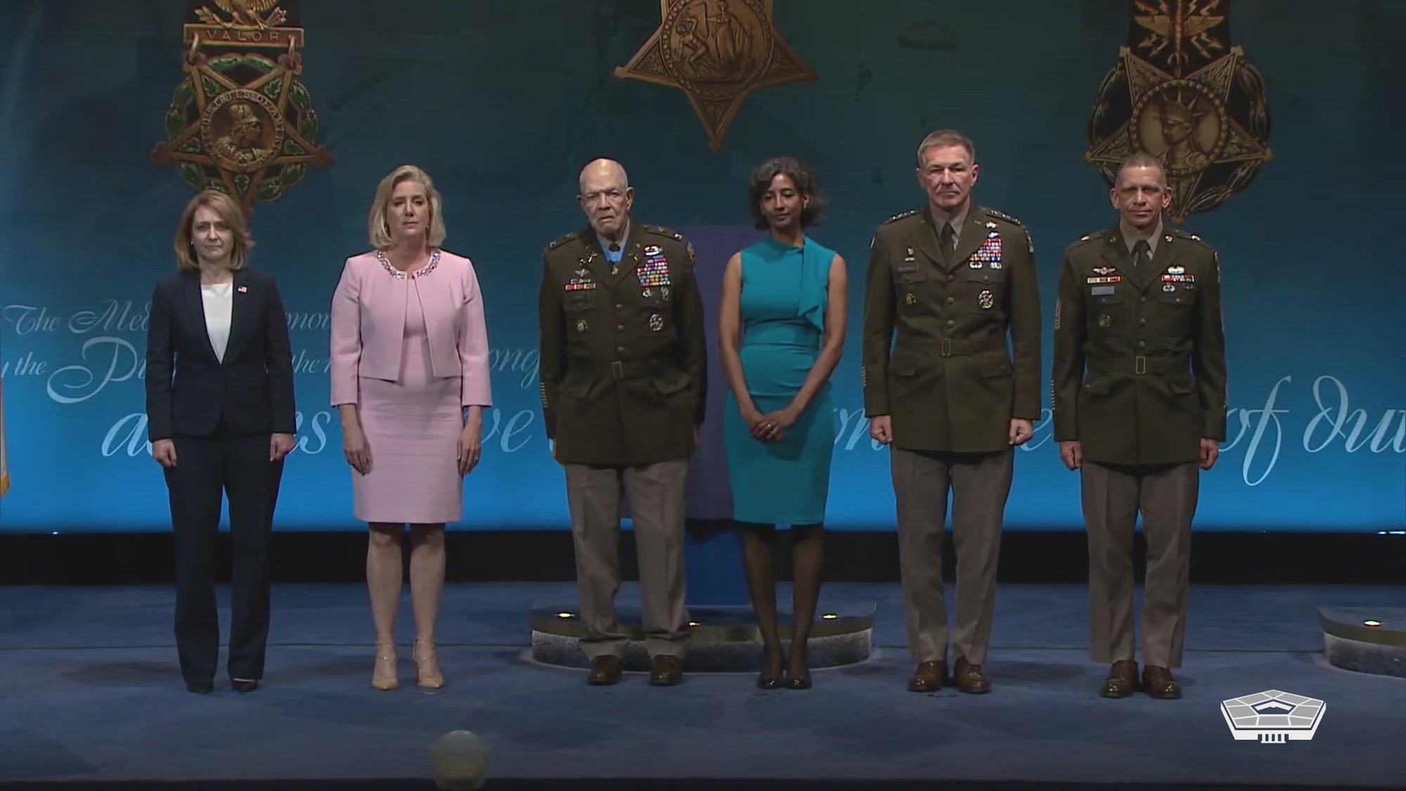 Six people, including three in Army uniform, stand in a row on a stage. 