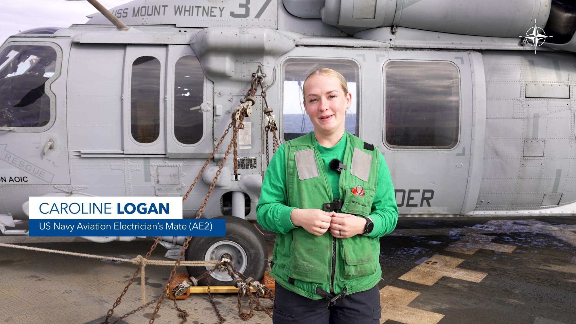 A sailor in a bright green vest stands in front of an aircraft on the deck of a ship.