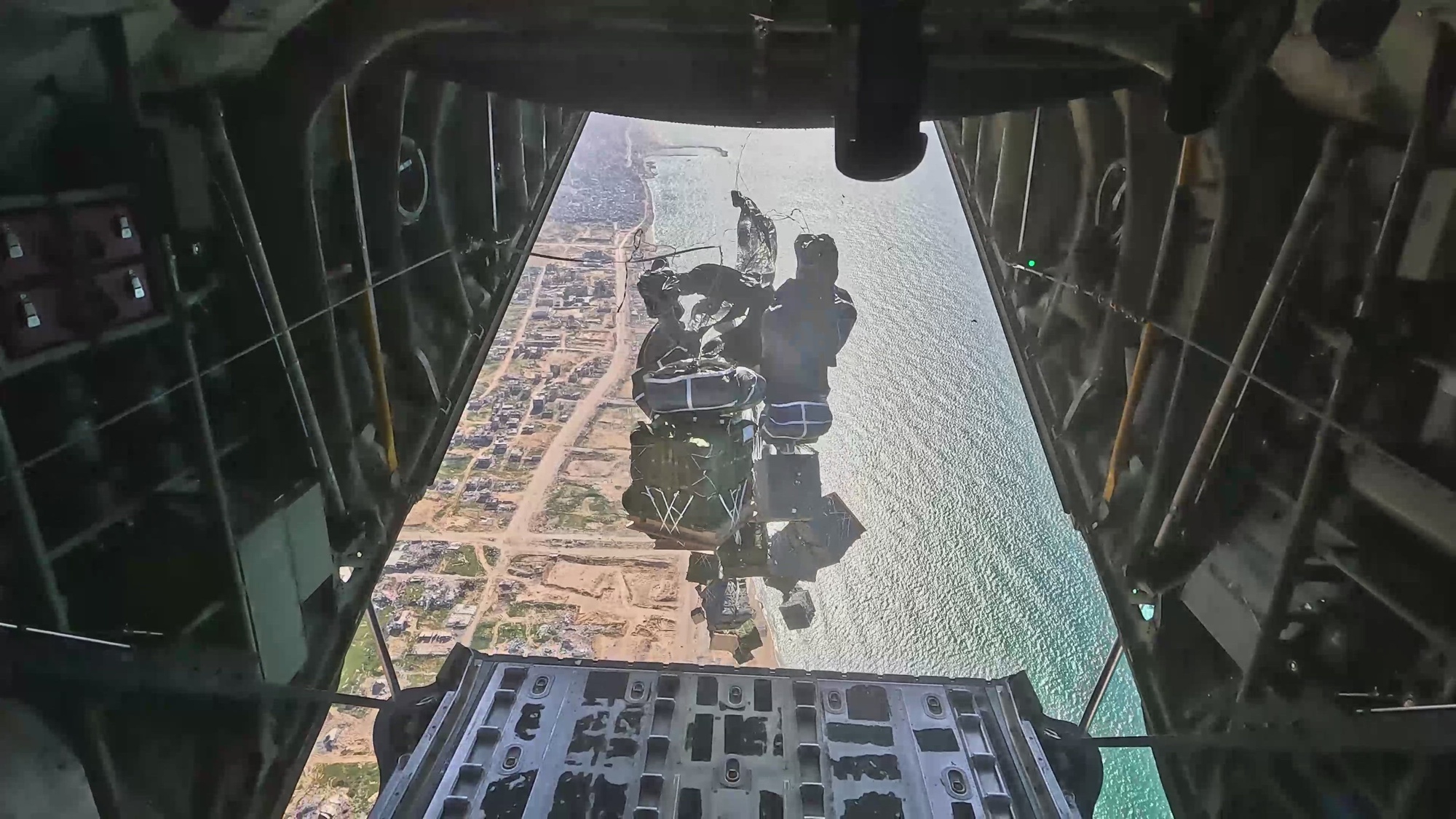 Pallets with parachutes attached fall from the back of an aircraft toward an area of land and water.