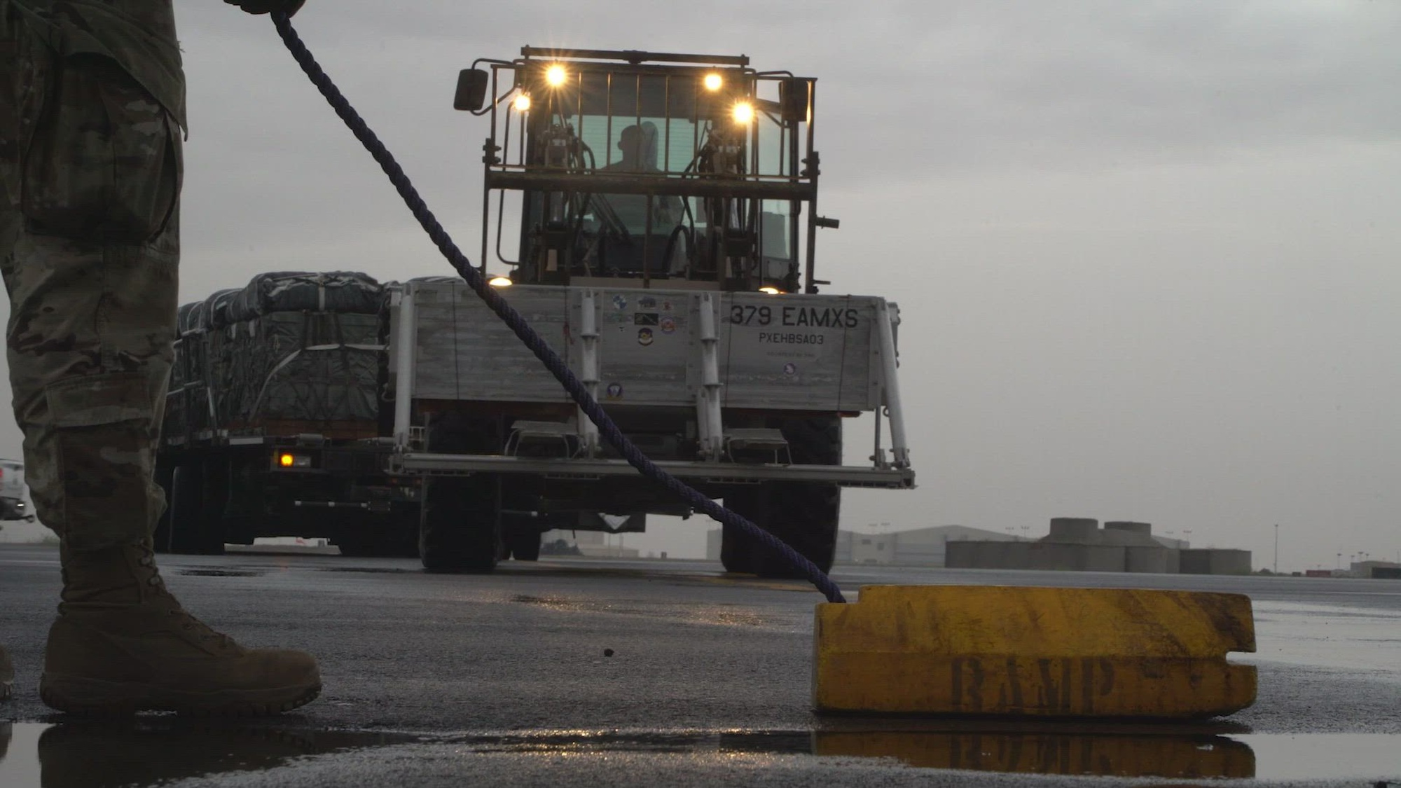 An airman operates a vehicle carrying pallets of humanitarian aid while another stands before it with a yellow tube on the ground.