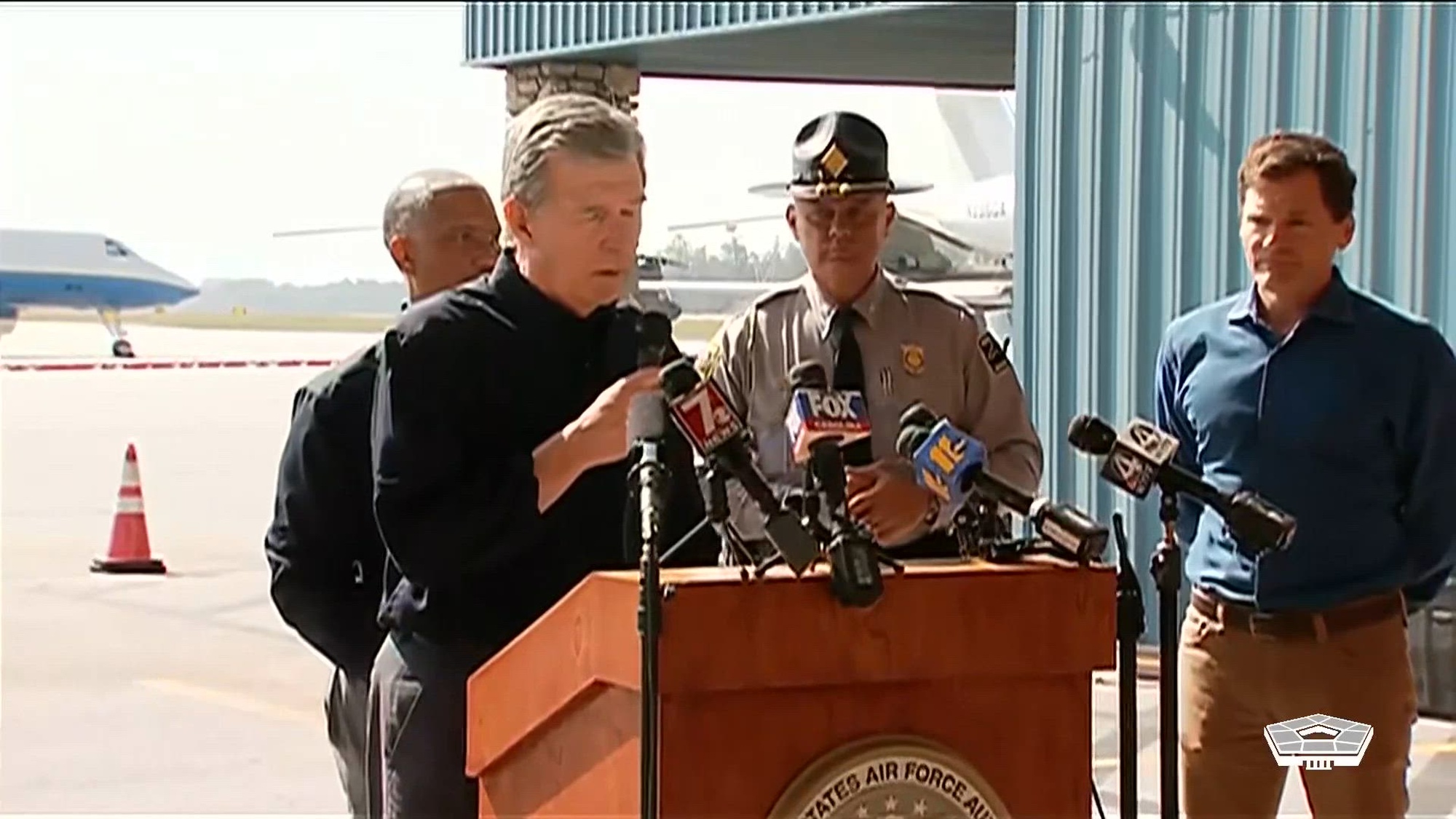 A civilian speaks at a lectern on a flight line as other officials stand nearby.