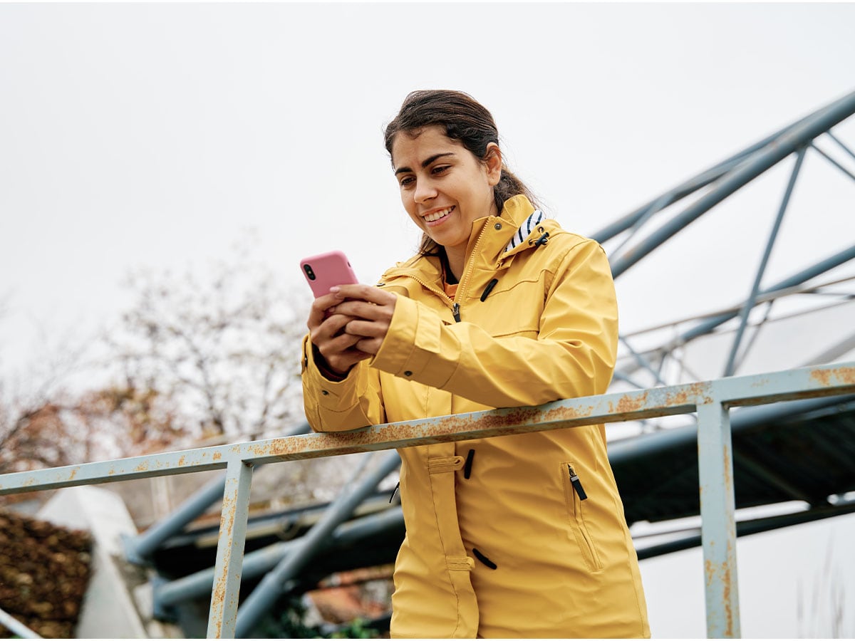 Wanita di jembatan memegang telepon mengenakan jaket kuning