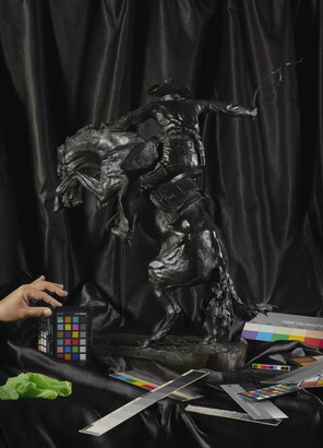 A color photograph of a bronze sculpture, from the back, of a cowboy on a rearing horse in front of black drapery as a light-skinned hand reaches in to hold up a color card. 