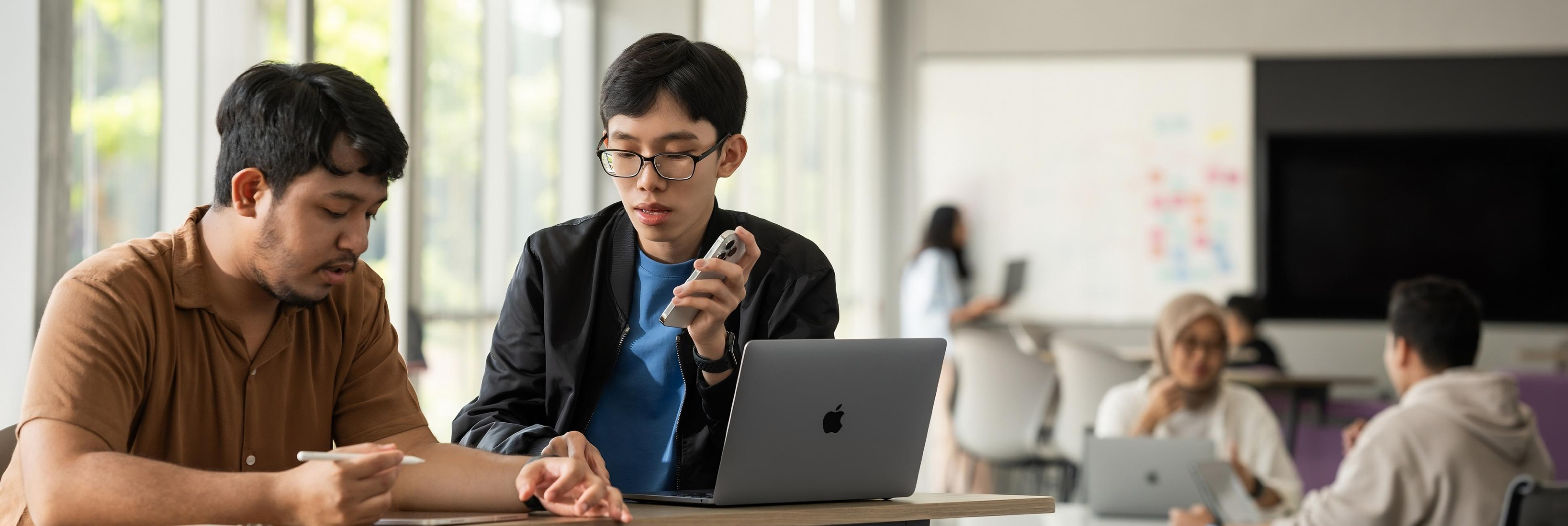 Jia Chen, an IT student and Apple Developer Academy mentor, collaborates with a colleague in an open workspace, using his iPhone and MacBook.