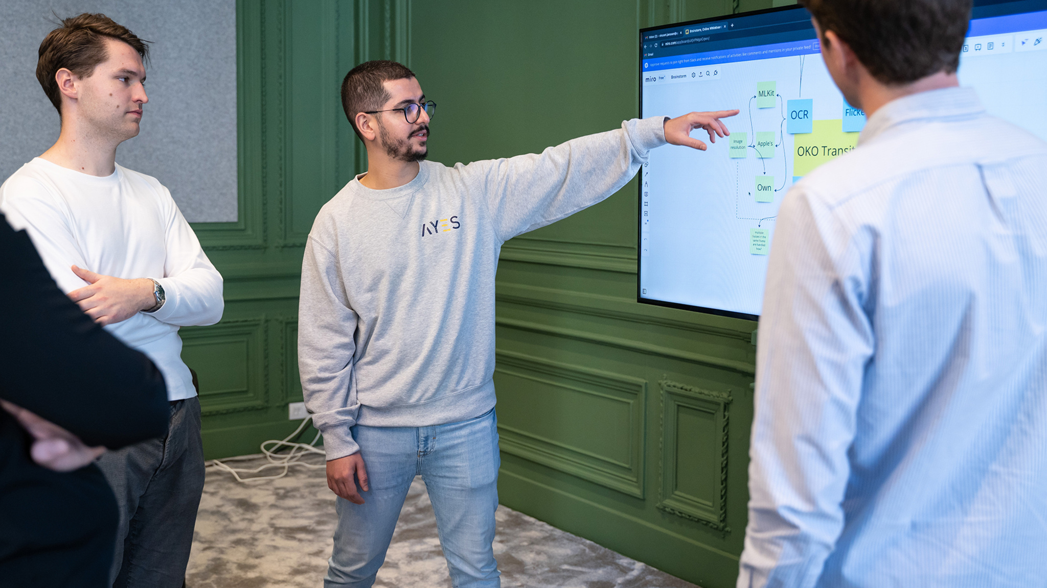 A group of men stand in a room with green walls looking at a large monitor that contains notes and sketches for the app *Oko*.