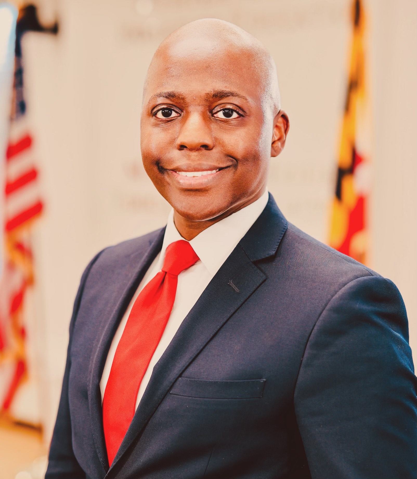 Dr. Erikk Bonner, smiling, wearing blue suite and standing in front of US and Maryland flags.