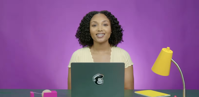 Woman with curly hair and yellow tshirt smiling at a desk facing an open laptop