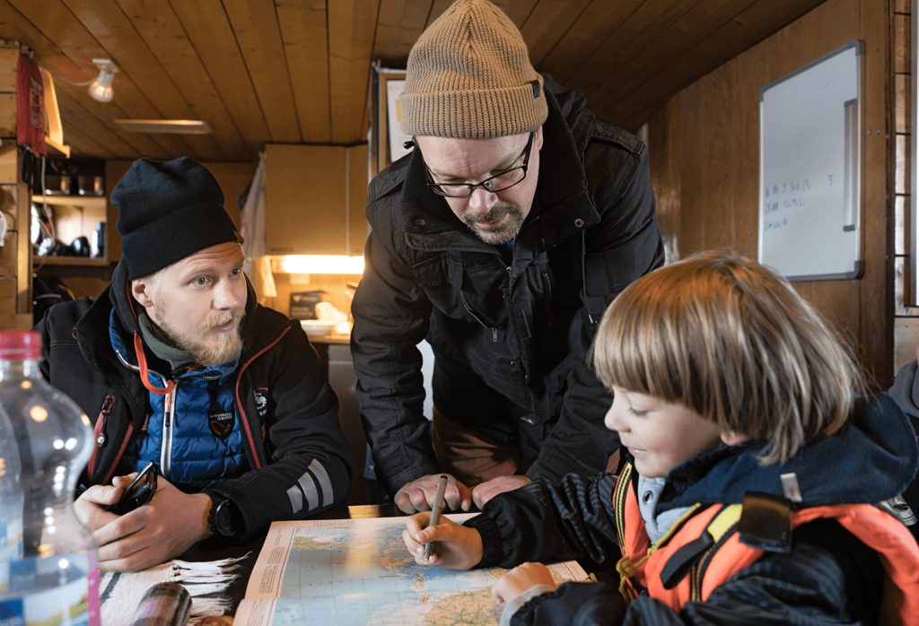 Kaksi miestä laivan sisätiloissa lukemassa merikorttia pikkupojan kanssa.