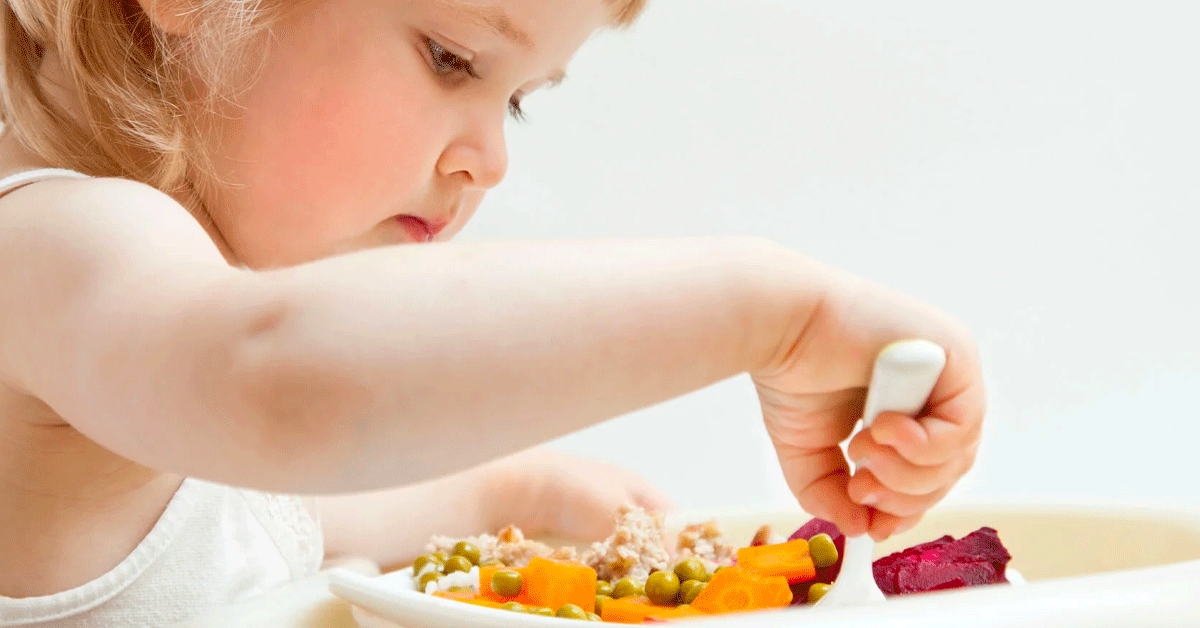 A blonde toddler holding a fork and eating the food on her plate.