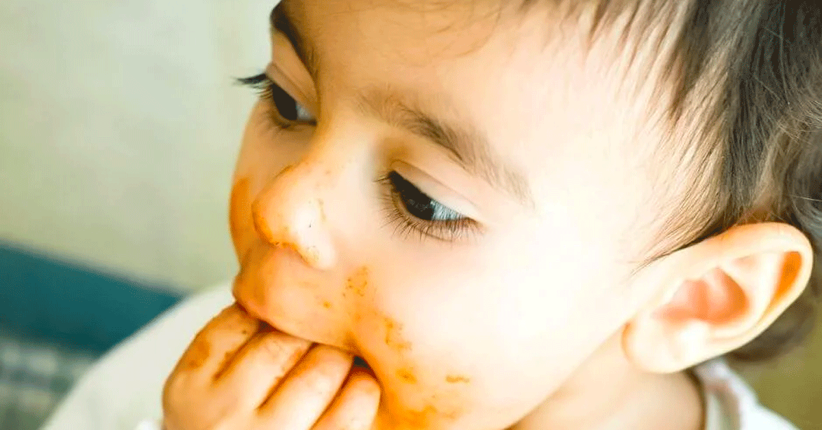 A toddler with food around her mouth and her hand and fingers in her mouth, while eating.