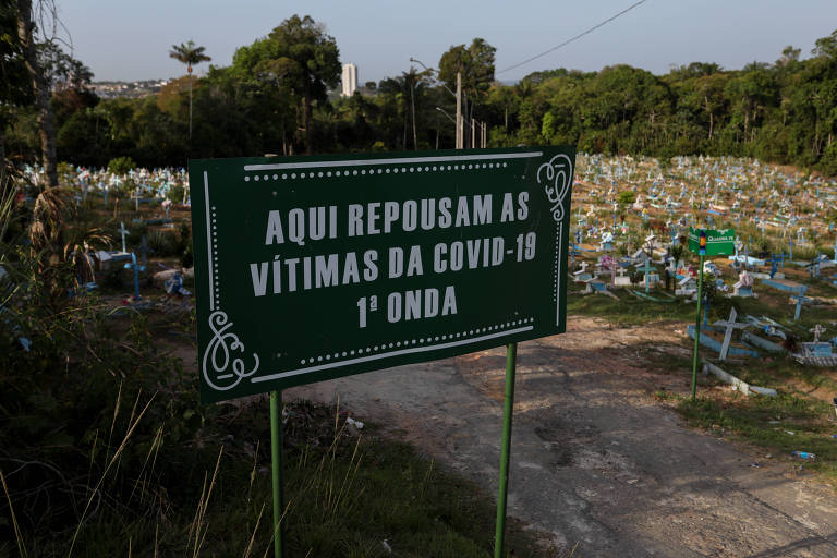 A imagem mostra um sinal verde em um cemitério, que diz: 'AQUI REPOUSAM AS VÍTIMAS DA COVID-19 1ª ONDA'. Ao fundo, há várias sepulturas cobertas com flores e grama, em um ambiente ao ar livre com árvores e vegetação ao redor.
