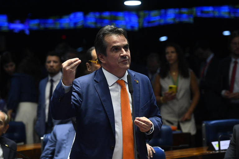 O senador está de pé, gesticulando. Ele usa terno azul, gravata laranja e camisa branca